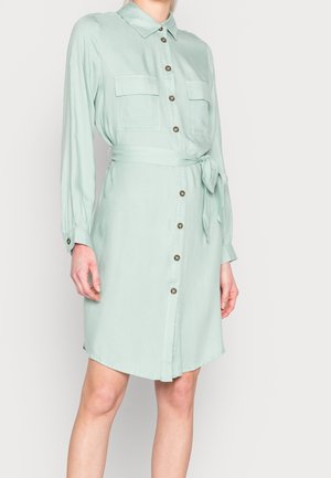 Femme portant une robe-chemise boutonnée vert clair avec manches longues, deux poches poitrine et un nœud à la taille, sur un fond uni.