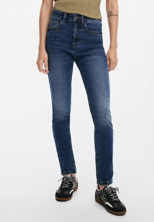 Person iført mørkeblå skinny jeans og sorte sneakers med hvide snørebånd, stående mod en ensartet lys baggrund.
