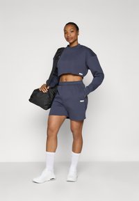 Marineblå cropped sweatshirt og matchende shorts lavet af blødt stof, med elastisk talje og lille mærkete. Hvide sneakers og sort taske.