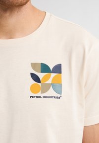 Krémové tričko s vícetrubicovým geometrickým logem složeným z kulatých tvarů v modré, žluté, zelené a béžové barvě.
