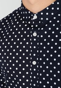 Marineblauwe shirt met witte stippen, voorzien van een button-up kraag en witte knopen langs de sluiting. Soepele, geweven textuur.
