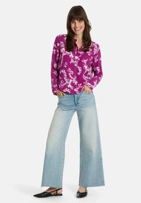 Langärmlige lila Bluse mit weißem Blumenmuster, kombiniert mit hellblauen weiten Jeans und schwarzen Slingbacks, aufrecht stehend.