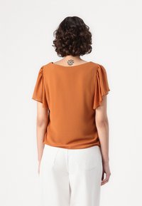 Blusa color caramelo con mangas cortas y acampanadas. Tejido suave, corte relajado, cuello redondo y espalda lisa. Combinada con pantalones de pierna ancha blancos.