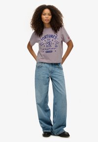 Camiseta gráfica morada con la frase "Fortunes Told" en azul, combinada con jeans de mezclilla azul claro holgados y zapatos negros.