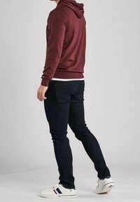 Sweat à capuche bordeaux avec cordon de serrage, associé à un jean slim bleu foncé et des baskets blanches avec des accents rouges et bleus.
