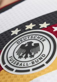 Emblème d'aigle noir avec texte "Deutscher Fussball-Bund" et quatre étoiles au-dessus sur un tissu texturé aux couleurs du drapeau allemand.