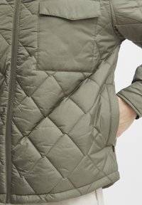 Quiltana maslinasto zelena jakna izrađena od sintetičkog materijala, s prednjim patent zatvaračem i zakrpantom na gornjoj lijevoj strani.