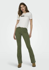 Camiseta blanca con el texto "EVOLUCIÓN", combinada con pantalones acampanados verdes y botas de tobillo marrones. Diseño sencillo, estilo casual.