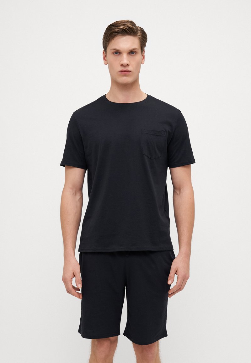 Schwarzes Baumwoll-T-Shirt mit kurzen Ärmeln und einer Brusttasche, kombiniert mit passenden schwarzen Shorts, die beide ein schlichtes, glattes Design aufweisen.