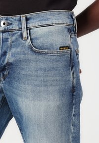 Lichtblauwe denim jeans met een vervaagde afwerking, voorzien van een metalen knoop, voorzakken en een zwart-geel logolabel op de achterzak.
