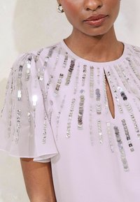 Light purple top with short sheer sleeves, featuring vertical rows of silver sequins and a keyhole neck detail. Smooth fabric texture.