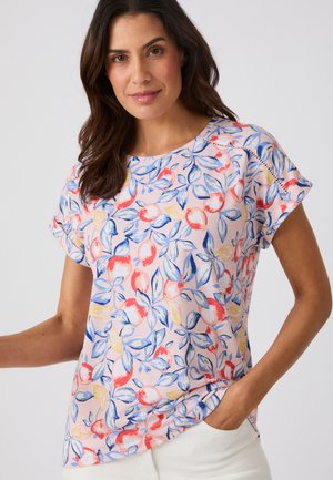 Frau mit dunklem Haar, die eine kurzärmelige, floral gemusterte Bluse mit blauen und roten Blättern trägt, posiert mit einer Hand in der Tasche einer weißen Hose.