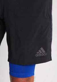 Svarta träningsshorts i lätt material, prydda med en diskret Adidas-logotyp, överlagda med blå kompressionsshorts.