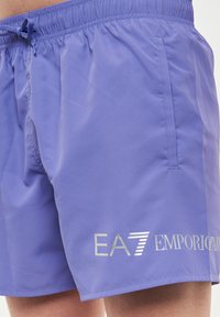 Pantaloni da bagno viola realizzati in tessuto leggero, con tasche laterali e una cintura elastica con cordino; logo EA7 ben in evidenza.
