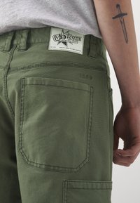 Olive green cargo pants with a woven label at the waistband, featuring back pocket stitching details and a textured fabric finish.