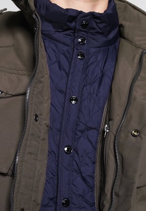 Gros plan d'une personne portant une veste matelassée bleu marine sous une veste vert olive avec fermeture éclair et boutons-pression.