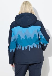 Osoba s blond vlasy, oblečená v námořnicky modré a modré bundě s motivem siluety hory a borovice, viděná zezadu.