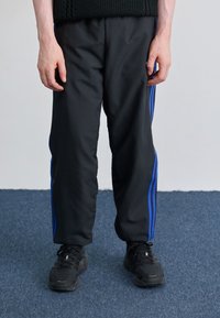 Sorte træningsbukser med blå sidelæg, lavet af et glat, mat stof, parret med sorte sneakers og står på blå tæppegulv.