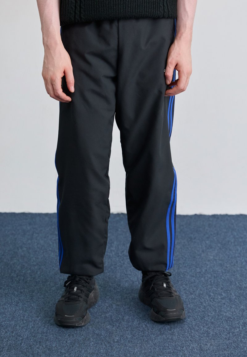 Sorte træningsbukser med blå sidelæg, lavet af et glat, mat stof, parret med sorte sneakers og står på blå tæppegulv.