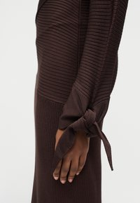 Σκούρο καφέ ριγωτό ένδυμα με μακριά μανίκια και κόμπο στο καρπό, φορεμένο στο χέρι ενός ατόμου πάνω σε απλό φόντο.