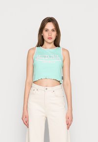 Top cropped verde menta com bainha ondulada, apresentando o texto branco "Von Dutch ORIGINALS". Usado com jeans off-white de cintura alta.