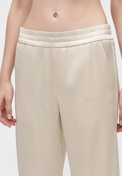 Gros plan sur un pantalon beige à taille élastique porté par une personne, montrant le bas du torse, les mains et un texte brodé subtil sur le côté droit.