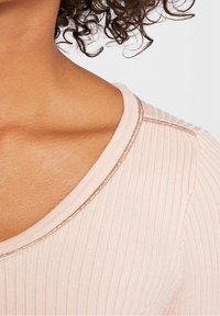 Kręcone pasma włosów opadające na ramię, ubrane w jasnoróżowy sweter w prążki z metalicznymi wykończeniami wzdłuż dekoltu i szwu na ramieniu.