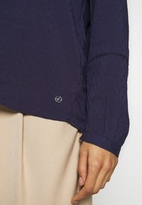 Blouse bleu marine texturée à manches longues, avec petits motifs brodés et un accent logo argenté près de l'ourlet.