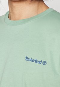 T-shirt vert menthe avec un col arrondi et des manches courtes. Présente un logo bleu "Timberland" et un graphique d'arbre sur le devant. Texture douce.