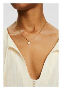 Collier en chaîne d'or avec un pendentif unique en perle ronde, porté sur une peau nue, accompagné d'une chemise en tissu texturé de couleur claire.