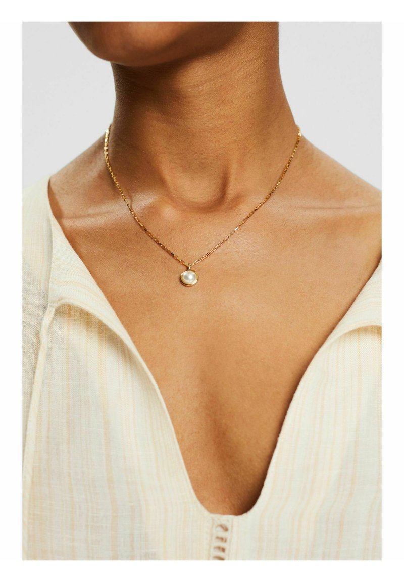 Collier en chaîne d'or avec un pendentif unique en perle ronde, porté sur une peau nue, accompagné d'une chemise en tissu texturé de couleur claire.