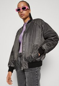 Giacca bomber nera con una finitura lucida e liscia. Presenta polsini e fondo a coste, chiusura con zip e tasche laterali. Indossata sopra un top viola.