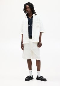 Set composto da una camicia bianca texturizzata e pantaloni corti abbinati, caratterizzato da una vestibilità rilassata. Sotto, una polo blu navy con scritte bianche. Scarpe nere e calze.