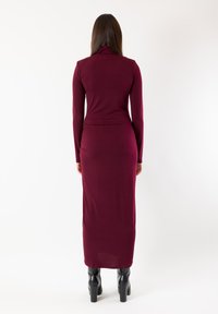 Abito bordeaux a maniche lunghe con silhouette aderente, collo alto e texture liscia, abbinato a stivaletti neri con plateau.