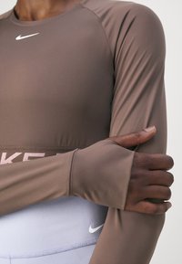Person, die ein braunes langärmliges Sportoberteil mit Daumenlöchern trägt und ihren Arm hält, sowie helllila Nike-Leggings mit sichtbaren Logos.