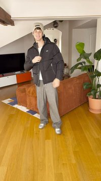 Zwarte donzen jas met ritszakken, grijze wijde broek, en zilveren sneakers, geplaatst in een binnenruimte met een bruine bank en planten.