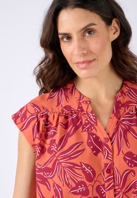 Blusa arancione con un design floreale in rosso scuro, dotata di maniche a ruffle corte, chiusura con bottoni e scollatura senza colletto.