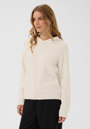 Femme aux cheveux blonds ondulés portant un pull à capuche blanc doux et un pantalon noir, debout devant un fond clair uni.