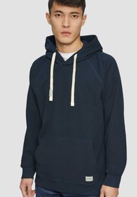Sudadera de un azul marino oscuro con un bolsillo tipo canguro, cordones de color crema, puños acanalados y una pequeña etiqueta en el dobladillo. Tejido suave y texturizado.