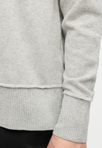 Κοντινό πλάνο ενός ατόμου που φοράει ανοιχτό γκρι πλεκτό πουλόβερ με ριμπ μανσέτες και τελείωμα, συνδυασμένο με μαύρο παντελόνι.