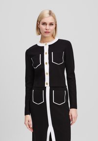 Black cardigan featuring white accents, two front pockets, and button closure. Long sleeves and cropped design emphasize a tailored look.