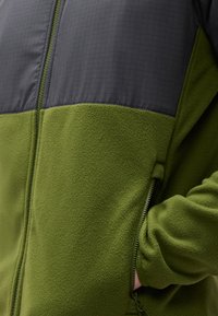 Πράσινο μπουφάν fleece με μαύρο πάνελ στο στήθος και ένα χέρι μερικώς μέσα σε μια πλαϊνή τσέπη με φερμουάρ.