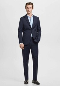 Mornarsko modro obleko iz teksturirane tkanine; enoredna jakna z odprtim ovratnikom, kombinirana s svetlo modro srajco z lisami in črnimi usnjenimi čevlji.