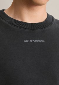 Zwarte sweatshirt met geribde halslijn en het logo "MARC O'POLO DENIM" in grijs, weergegeven in een zachte, gestructureerde stof. Close-up van het kraaggebied.