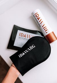 Svart stoff selvbruningsapplikatorhanske med hvit "IDAWARG" logo, ved siden av en hvit og kobber selvbruningsspray og en svart applikatorpose.