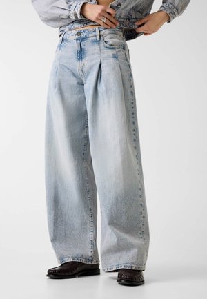 Personne portant un jean large bleu clair avec des plis, des mocassins foncés et une veste en denim assortie, ajustant les boutons avant de la veste.