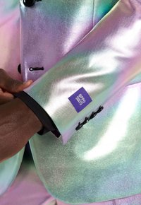 Manica di un abito iridescente brillante con bottoni neri e etichetta "Opposuits", indossata sopra un polsino nero su un braccio di carnagione scura.