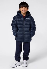 Giacca a imbottitura blu navy con cappuccio, caratterizzata da un design trapuntato e chiusura con zip. Indossata sopra pantaloni scuri e sneakers bianche. Logo sul petto.