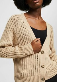 Beige gestrickte Strickjacke mit V-Ausschnitt, Knopfleiste und gerippten Bündchen. Verfügt über ein strukturiertes Muster mit offenen Maschen und entspannter Passform.