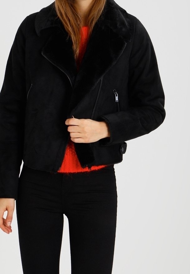 Czarna kurtka z sztucznego zamszu, krótka, z szerokim kołnierzem, kieszeniami na zamek błyskawiczny i miękką podszewką, noszona na czerwonym swetrze i dopasowanych czarnych spodniach.